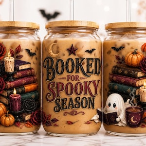 May include: Three glass tumblers with bamboo lids and straws, filled with iced coffee. The design features stacks of books, pumpkins, lit candles, roses, bats, and a ghost. The center tumbler displays the text "BOOKED FOR SPOOKY SEASON" in a decorative font.