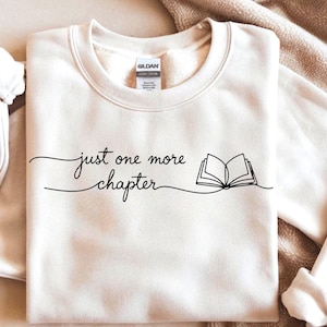 Puede incluir: Sudadera color crema con la frase "just one more chapter" en fuente manuscrita, acompañada de un dibujo lineal de un libro abierto. La sudadera está hecha de un material suave y es adecuada para uso casual.