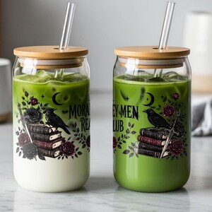 Two glass tumblers with straws, one white drink, one green, decorated with raven and book illustrations.