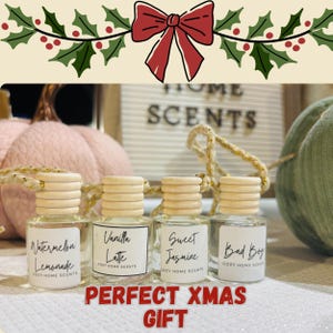 May include: Four small glass bottles with wooden caps, labeled with scents like "Watermelon Lemonade" and "Sweet Jasmine." The bottles are arranged in front of a sign that says "HOME SCENTS." The text "PERFECT XMAS GIFT" is at the bottom.