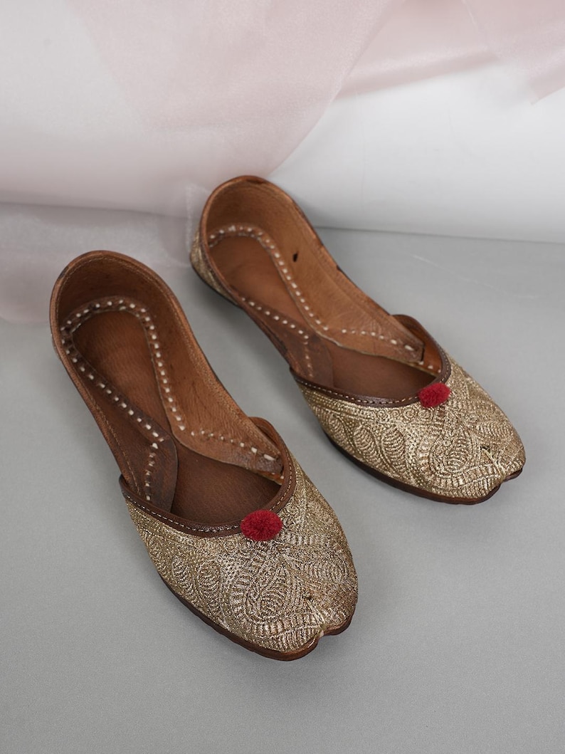 Pode incluir: Um par de sapatilhas bordadas douradas com interior e acabamento em couro castanho. Cada sapato apresenta um pequeno detalhe de pompom vermelho na ponta. As sapatilhas t&ecirc;m um design tradicional.