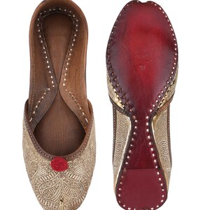 Pode incluir: Par de sapatos slip-on bordados dourados com um pompom vermelho e interior em couro castanho. A sola &eacute; vermelha com detalhes dourados. Os sapatos s&atilde;o exibidos em um fundo branco.