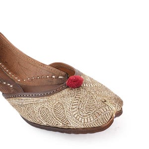 Pode incluir: Close-up de um sapato bordado dourado com um detalhe de pompom vermelho. O sapato apresenta padr&otilde;es intrincados e uma sola de couro castanho. O design &eacute; detalhado e o sapato parece ser feito &agrave; m&atilde;o.