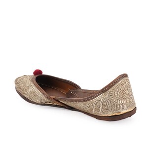 Pode incluir: Sapato raso dourado com bordados intrincados e um pequeno pompom vermelho. O sapato tem sola e acabamento em couro castanho, e um padr&atilde;o decorativo cobre a parte superior.
