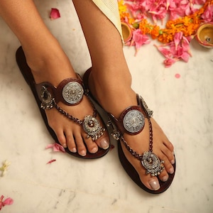 Könnte beinhalten: Braune Ledersandalen mit silbernen Verzierungen. Die Sandalen haben ein kreisförmiges silbernes Medaillon am oberen Riemen und eine Kette mit Anhänger. Die Sandalen sind im Zehensteg-Stil.