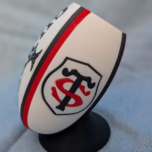 Peut inclure: Une réplique de ballon de rugby blanc avec une bande rouge et noire, un socle circulaire noir et un emblème en forme de bouclier arborant un monogramme stylisé noir et rouge des lettres T et S. La surface présente une finition texturée.