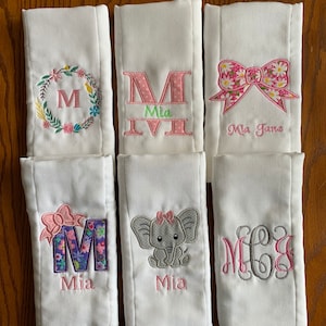 May include: Six white burp cloths with embroidered designs. The designs include a floral wreath with the letter M, a pink and white polka dot M with the name Mia, a pink and white bow with the name Mia Jane, a floral M with the name Mia, a gray elephant with a pink bow and the name Mia, and a gray monogram with the letters MCG.