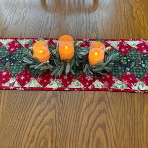 May include: A festive Christmas table runner with a patchwork design in red, green, and black. Three lit candles in glass holders are centered on the runner, surrounded by greenery. The runner is placed on a wooden table.