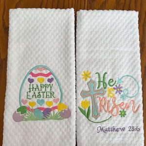 May include: Two white kitchen towels with embroidered Easter designs. One towel features a colorful Easter egg with the text "Happy Easter". The other towel features a cross with the text "He is Risen" and the scripture reference "Matthew 28:6".