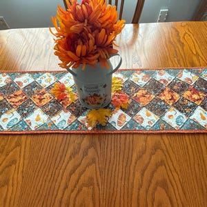 May include: A fall-themed table runner with a patchwork design featuring pumpkins and leaves in shades of orange, brown, and teal. A small, light blue pitcher with orange flowers sits on top of the runner, adding a decorative touch.