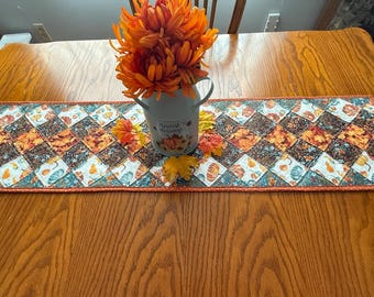May include: A fall-themed table runner with a patchwork design featuring pumpkins and leaves in shades of orange, brown, and teal. A small, light blue pitcher with orange flowers sits on top of the runner, adding a decorative touch.