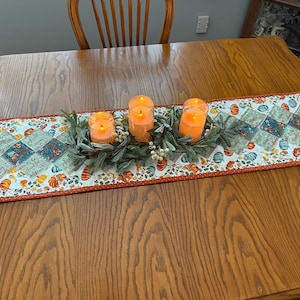 May include: A decorative table runner with a fall theme. The runner features a patchwork design with pumpkins and floral patterns in orange, teal, and cream colors. Three orange candles in glass holders sit on top of the runner, surrounded by greenery.