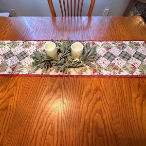 May include: A quilted table runner with a patchwork design featuring red, green, and white squares. Two battery-operated candles sit atop a decorative greenery arrangement. The runner has a red border and is placed on a wooden table.