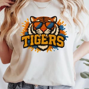 May include: Cream-colored t-shirt featuring a tiger wearing sunglasses, with the word "TIGERS" in a bold, yellow font. The tiger design is surrounded by orange and blue accents, creating a vibrant and eye-catching graphic.