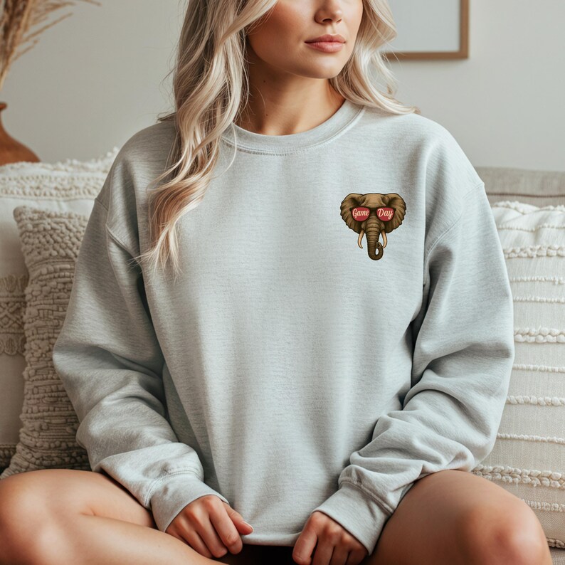 May include: Light gray sweatshirt featuring a cartoon elephant wearing sunglasses with the words "Game Day" on the lenses. The elephant graphic is brown with ivory tusks. The sweatshirt is a crew neck style.