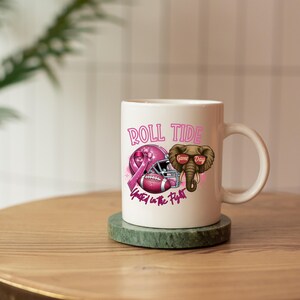 May include: White ceramic mug with a pink football helmet, a pink ribbon, and an elephant wearing sunglasses. The mug has the words "Roll Tide" and "Game Day" printed on it. The phrase "United in the Fight" is also printed on the mug.