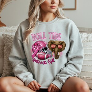 May include: Light gray sweatshirt with a graphic design featuring a pink football helmet, football, and an elephant wearing sunglasses. The text "ROLL TIDE" and "United in the Fight" are also displayed in pink.