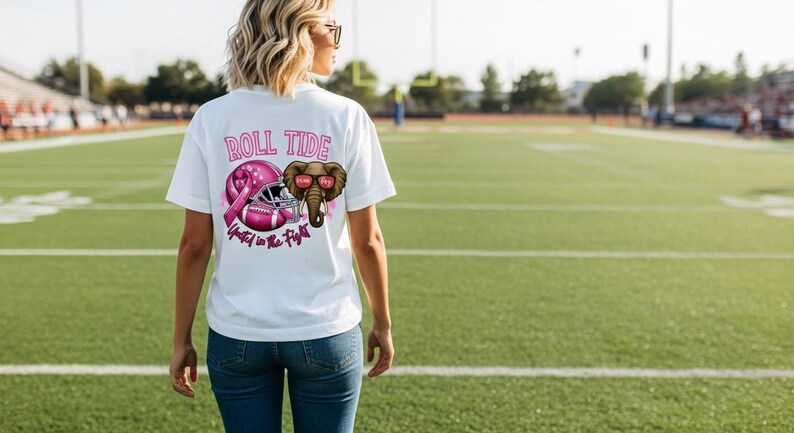 May include: White t-shirt with a pink graphic featuring a football helmet, a pink ribbon, and an elephant wearing sunglasses. The text "ROLL TIDE" and "United in the Fight" are also printed on the shirt.