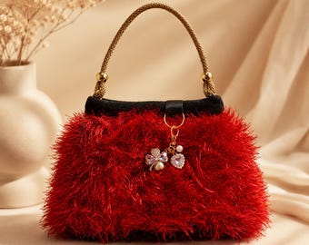 May include: A red, fluffy handbag with a black top and gold handle. The bag has a decorative charm with clover, heart, and pearl accents. The handle is a twisted gold rope.