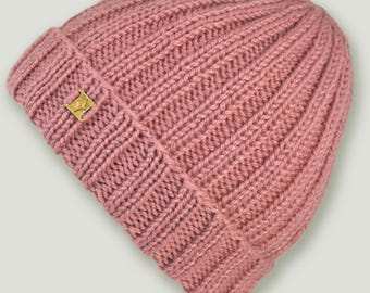 May include: A dusky pink ribbed knit beanie hat. The hat has a small, gold-coloured square emblem on the folded brim. The beanie is made of a thick, warm material, suitable for cold weather.