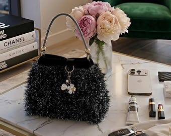 May include: A black handbag with a fluffy, textured exterior and a beaded handle sits on a marble table. The bag has a decorative charm and is next to a bouquet of pink and white flowers, a phone, and cosmetics.