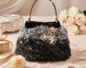 May include: A small, fluffy handbag with a black and grey ombre design. The handle is embellished with sparkling rhinestones and gold accents. A charm with a heart and clover hangs from the handle. The bag is set on a pink surface with roses and a gift box.