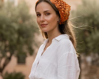 May include: An orange crochet headscarf with an openwork pattern worn over long hair, paired with a white button-down shirt and light blue denim jeans. The headscarf features a textured, knotted yarn construction that covers the top of the head.