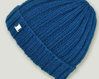 May include: A blue knitted beanie hat with a ribbed design. The hat has a small, silver fox emblem on the folded brim. The beanie is made of a thick, warm material, suitable for cold weather. The hat is a solid blue colour.