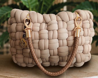 May include: A beige handbag constructed from thick, woven fabric tubes in a grid pattern. It features a single, braided brown handle attached with gold-toned metal clasps. A small, circular gold-toned charm hangs from one of the side attachment rings.