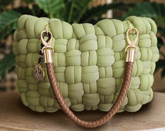 May include: A light green handbag constructed from thick, woven fabric strips in a puffed, grid-like pattern. It features a single, braided brown handle attached with gold-toned metal clasps and a small, circular metallic charm hanging from one side.