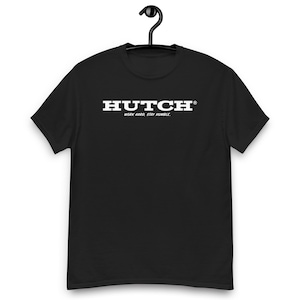 Peut inclure: T-shirt noir avec le texte blanc "HUTCH" et le slogan "WORK HARD. STAY HUMBLE." Le t-shirt est suspendu à un cintre noir. Le t-shirt est de style col rond classique.