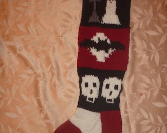 May include: A red, white, and black knitted stocking with a ghost, a bat, and two skulls. The stocking has the name "Nick" knitted into the top.