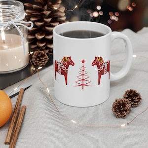 May include: White ceramic mug featuring two red Dala horses with decorative patterns and a red Christmas tree design. The mug is filled with a dark liquid and has a large handle. The mug is surrounded by festive decorations.