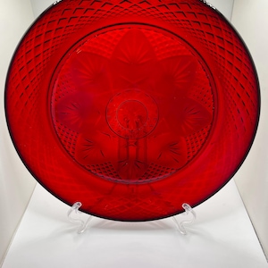 May include: A vibrant red glass plate with a diamond pattern around the rim and a floral design in the center. The plate is round and sits on a clear plastic stand, showcasing its intricate details and rich color.