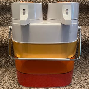 May include: A vintage, two-tiered lunch pail with a white top, a yellow middle section, and a red base. It has a metal handle and two white, cylindrical compartments on top with orange accents and white latches.
