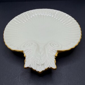 May include: A white ceramic dish shaped like a scallop shell with a glossy finish and a gold-painted rim. The surface features embossed radial ridges extending from the base, which is decorated with a raised, symmetrical leaf-like pattern.