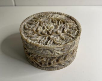 May include: A round, beige decorative box with intricate carved floral patterns. The box is cylindrical with a lid and base, and the carvings cover the entire surface. The box is made of a light-colored material, possibly stone or ceramic.