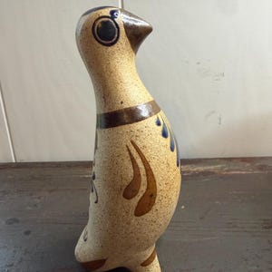 May include: A ceramic bird figurine with a speckled beige finish, featuring hand-painted brown and blue decorative accents. The bird has a rounded body, a pointed beak, and a dark brown band around its neck, standing on a small base.