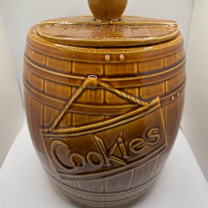 May include: A brown ceramic cookie jar with a barrel design. The jar has a lid with a round knob and a sign that reads "Cookies". The jar is a warm brown color.