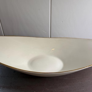 May include: Cream-colored, elongated ceramic bowl with a gold rim. The bowl has a shallow, oval shape with a slightly raised center. The gold trim adds a touch of elegance to this decorative piece, suitable for serving or display.