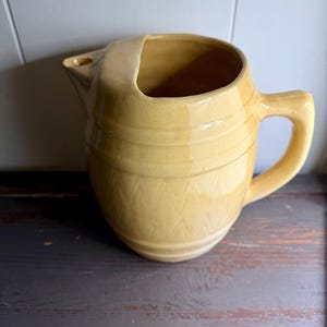 May include: A mustard yellow ceramic pitcher with a glossy finish, featuring a wide spout, a sturdy side handle, and a textured pattern of horizontal bands and subtle geometric indentations around the rounded body of the vessel.