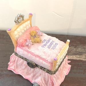 May include: A miniature bed-shaped trinket box with a pink comforter and a teddy bear. The bed frame is light brown with purple accents. The text "THE PRINCESS SLEEPS HERE" is on the comforter. A small gray cat peeks out from under the bed.