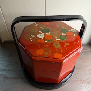 May include: A three-tiered octagonal red lacquer food container with a black handle and base. The lid features a decorative gold, green, and orange floral pattern of plum blossoms. The stackable compartments are uniform in shape and color.