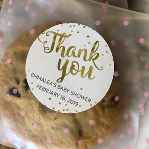 Op de afbeelding: Een witte ronde sticker met gouden letters die "Bedankt" en "Emmalea's Baby Shower February 16, 2019" zeggen. De sticker zit op een doorzichtige plastic zak met roze stippen en een chocoladekoekje erin.
