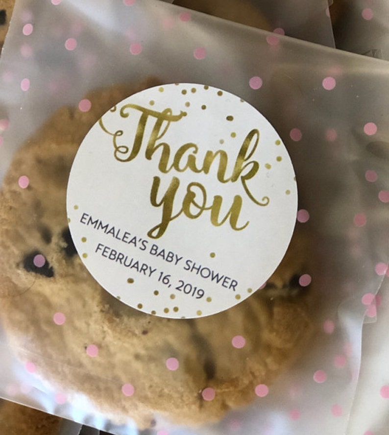 Op de afbeelding: Een witte ronde sticker met gouden letters die "Bedankt" en "Emmalea's Baby Shower February 16, 2019" zeggen. De sticker zit op een doorzichtige plastic zak met roze stippen en een chocoladekoekje erin.