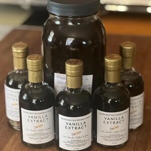 May include: Five glass bottles of vanilla extract, with gold lids and labels that say "Handcrafted Double-Fold Vanilla Extract". The bottles are arranged on a wooden surface, with a larger glass jar of vanilla extract in the background.
