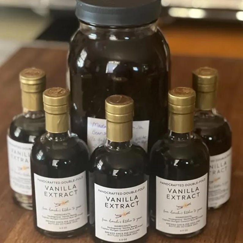 May include: Five glass bottles of vanilla extract, with gold lids and labels that say "Handcrafted Double-Fold Vanilla Extract". The bottles are arranged on a wooden surface, with a larger glass jar of vanilla extract in the background.