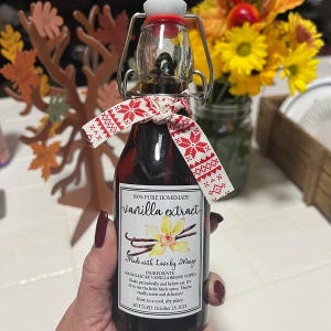 May include: A small glass bottle of homemade vanilla extract with a decorative red and white ribbon. The label reads "100% Pure Homemade Vanilla Extract" and includes the text "Made with Love by Marge."