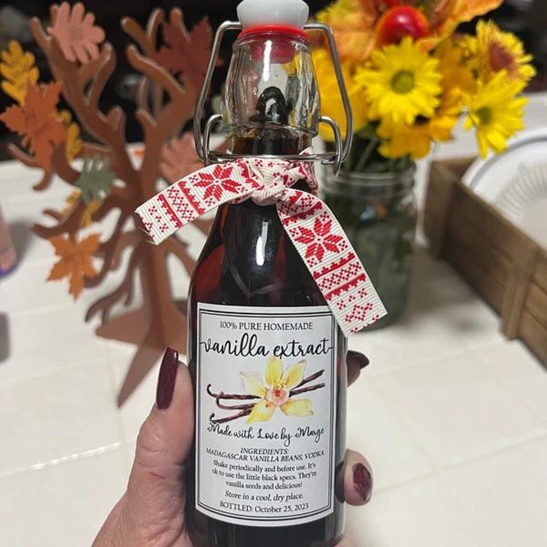 May include: A small glass bottle of homemade vanilla extract with a decorative red and white ribbon. The label reads "100% Pure Homemade Vanilla Extract" and includes the text "Made with Love by Marge."