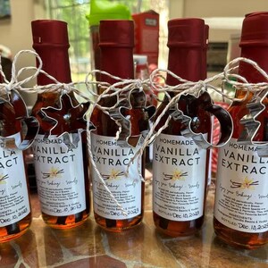May include: Five small glass bottles filled with brown liquid and labeled "Homemade Vanilla Extract". Each bottle is decorated with a white ribbon and a metal cookie cutter in the shape of a star. The bottles are arranged in a row on a brown surface.